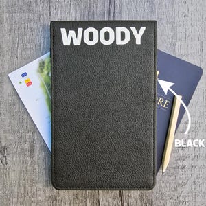 Könnte beinhalten: Schwarzes Leder-Notizbuch mit dem Namen "WOODY" in weißen Blockbuchstaben. Ein Bleistift und ein blaues Notizbuch sind darin verstaut. Auf der linken Seite ist eine kleine Karte zu sehen.
