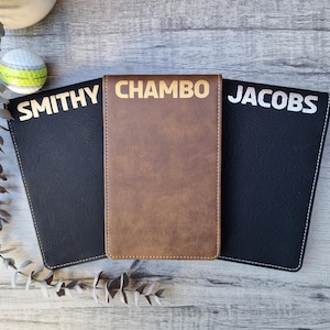 May include: Three personalized golf score card holders. One is brown with "CHAMBO" in gold lettering. The other two are black with white stitching and names "SMITHY" and "JACOBS" in silver lettering. A golf ball is visible in the background.