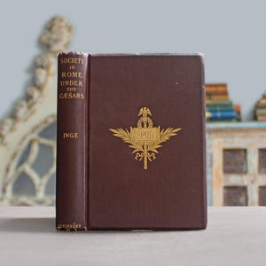 May include: A dark brown hardcover book titled Society in Rome Under the Caesars by Inge, published by Scribners. The front cover features a gold embossed emblem of a staff with wings and the letters SPQR centered on the book cover.