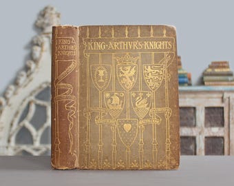 May include: A vintage hardcover book titled King Arthur's Knights by Henry Gilbert, illustrated by Walter Crane. The brown cover features gold-embossed illustrations of six heraldic shields, a crown, and vertical spear-like lines throughout.