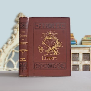 May include: A maroon-coloured hardcover book titled "The Story of Liberty" with gold lettering and illustrations. The cover features a design with a torch, olive branch, and other symbols. The spine displays the author's name and publisher.