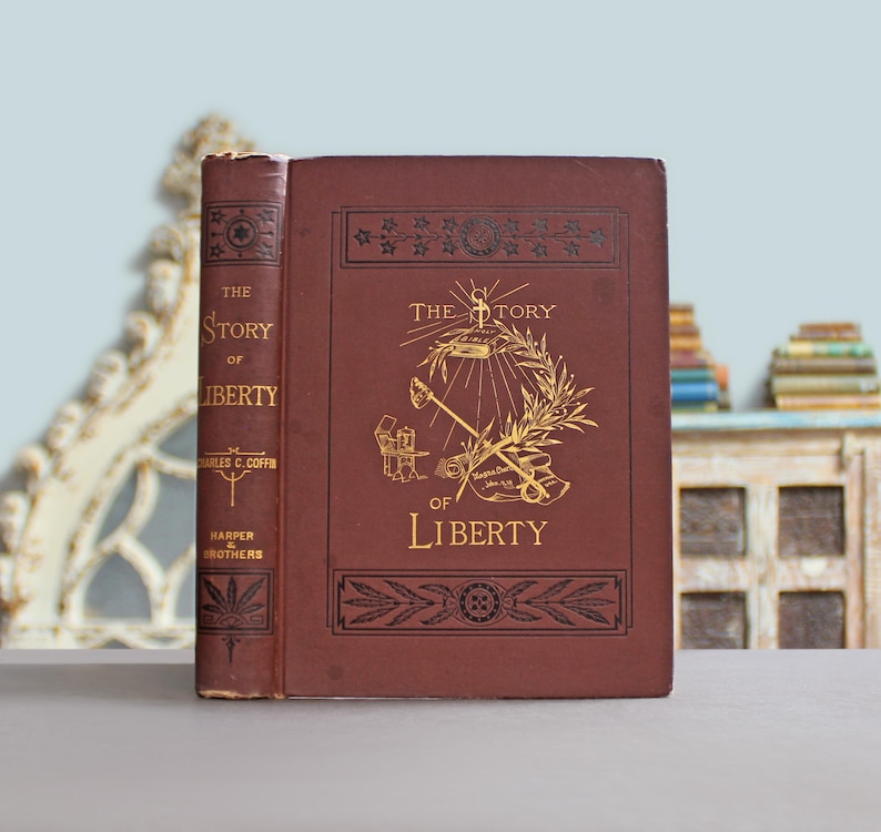 May include: A maroon-coloured hardcover book titled "The Story of Liberty" with gold lettering and illustrations. The cover features a design with a torch, olive branch, and other symbols. The spine displays the author's name and publisher.
