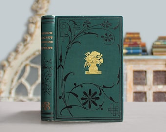 Op de afbeelding: Een vintage boek met een donkergroene kaft met gouden bloemaccenten en een gouden vaas met bloemen. De rug toont de titel "WOOD'S OBJECT LESSONS IN BOTANY" in gouden letters. Het boek heeft een decoratief bloemenontwerp.