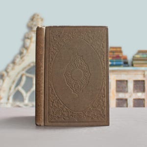 May include: A vintage book with a brown cover featuring an embossed oval design. The book's spine is a lighter shade of brown, and the pages are slightly aged. The book is standing upright on a neutral surface.