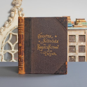 May include: A vintage book titled "Character Sketches of Romance Fiction and the Drama" with gold lettering on a brown cover. The spine is a lighter brown with text. The book is on a gray surface.