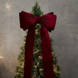 May include: A Christmas tree adorned with a large, deep red velvet bow. The bow is the focal point, with long, flowing tails. The tree is lit with warm white lights, creating a festive holiday display.