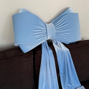 May include: A large, light blue velvet bow, possibly a decorative pillow, rests on a dark brown sofa. The bow has a detailed, textured design with long, flowing tails. The soft, plush material and color create a visually appealing accent piece.