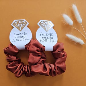 May include: Two rust-colored satin scrunchies with white tags that say "I can't tie the knot without you" and feature a gold diamond ring illustration.