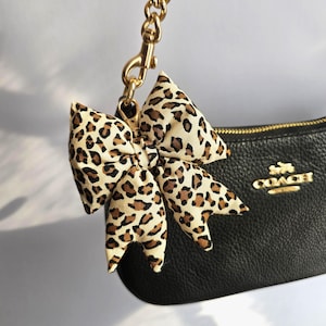 May include: A leopard print bow charm attached to a gold chain and clasp. The bow is made of a cream-coloured fabric with brown spots. The charm is attached to a black leather Coach purse with a gold zip and logo.