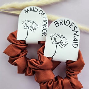 May include: Two rust-colored satin scrunchies with white tags. The tag on the left says "Maid of Honor" and the tag on the right says "Bridesmaid".