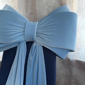 May include: A light blue velvet bow chair cover. The bow is large and decorative, with a central knot and flowing fabric. The chair itself is a dark blue color, and the background features a floral patterned curtain.
