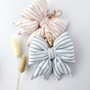 May include: Two fabric bows with a striped pattern, one in pink and white and the other in blue and white. Each bow features a metallic gold-toned lobster clasp attached to the centre knot, allowing for easy attachment to bags or accessories.
