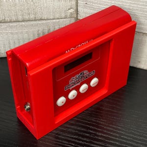 May include: A bright red electronic device with the text "MD-Cross 10" and "Decimator Design" visible. It has a small screen and several white buttons. The device is rectangular and appears to be a piece of audio or video equipment.