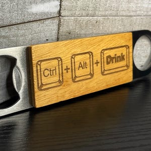 May include: A bottle opener with a stainless steel head and a wooden handle. The handle has the words "Ctrl + Alt + Drink" engraved on it. The opener is designed for opening bottles.