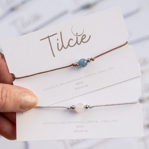 May include: Two handmade crystal bracelets on white cards. The top bracelet has a light blue stone and brown string. The bottom bracelet has a light pink stone and a grey string. Both cards have the text "Tilcie Crystals and Handmade Crystal Jewellery @tilcie_ tilcie.com.au"