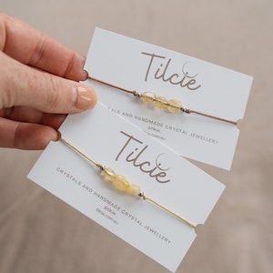 May include: Two handmade crystal bracelets on white cards. Each bracelet features yellow-orange gemstones and silver beads strung on a cord. The cards display the brand name "Tilcie" and the text "CRYSTALS AND HANDMADE CRYSTAL JEWELLERY".