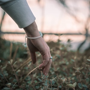 Peut inclure: Une main portant un bracelet en cordon blanc avec des perles turquoise tend la main pour toucher une plante verte.