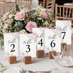 May include: White table number cards with floral accents and black numerals 2, 3, 4, 6, and 7. Each card is held by a small, square wooden block. A bouquet of pink and white roses sits behind the cards, with wine glasses and plates in the foreground.