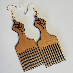 May include: Wooden earrings featuring a fist design atop a comb shape. The earrings are light brown with black outlining and gold-colored ear wires. The design is a symbol of solidarity and empowerment.