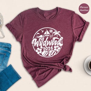 a casual outfit consisting of a burgundy t-shirt with a graphic design, a pair of blue jeans, a pair of black sneakers, a pair of glasses, a white coffee cup, and a straw hat.