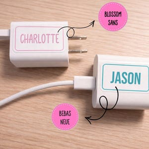 May include: Two white phone chargers with personalized labels. One charger has "CHARLOTTE" in pink, the other has "JASON" in teal. The chargers are connected to white cords and sit on a light wood surface.
