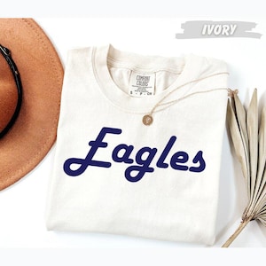 May include: An ivory short-sleeve t-shirt with a crew neckline featuring the word Eagles printed in a dark blue cursive script across the chest. The shirt has a Comfort Colors brand label on the inside collar and is size S.
