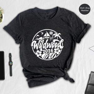 a casual outfit laid out on a white surface, including a black t-shirt with a graphic design, a tablet, a watch, a potted plant, and a black bag.