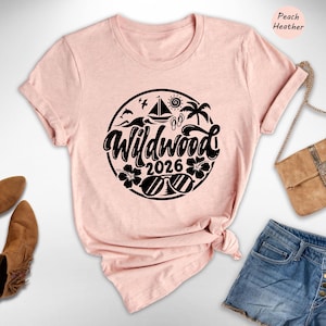 A pink t-shirt with a graphic design, a pair of brown ankle boots, a beige handbag, and a pair of denim shorts are arranged on a white background.