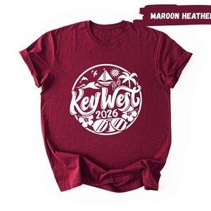 A maroon t-shirt with a white graphic design featuring the text "Key West" and "2016" surrounded by various beach and ocean-themed illustrations.