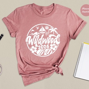 a pink t-shirt with a white graphic design, a watch, a notebook, a pen, and a pink handbag arranged on a beige background.