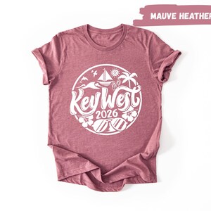 A maroon t-shirt with a white graphic design featuring the text "Key West" and "2016" surrounded by various beach and ocean-themed illustrations.