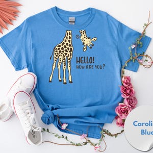 A blue t-shirt with a giraffe and the text "HELLO HOW ARE YOU?" printed on it, along with a pair of white sneakers and pink flowers.