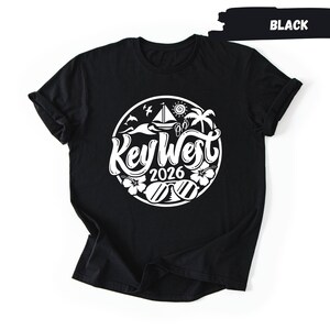 A black t-shirt with a white graphic design on it. The design features a circular motif with various elements such as a sailboat, palm trees, and a bird. The text "Key West" is prominently displayed in the center of the design.