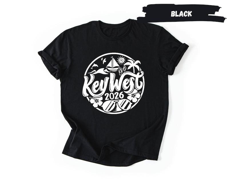A black t-shirt with a white graphic design on it. The design features a circular motif with various elements such as a sailboat, palm trees, and a bird. The text "Key West" is prominently displayed in the center of the design.