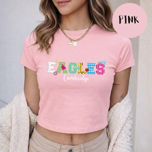 May include: A light pink short-sleeved t-shirt featuring the word EAGLES in colorful, patterned block letters with a small flower and apple icon. Below the word, the text Cambridge is printed in a white script font. A gold chain necklace is worn.