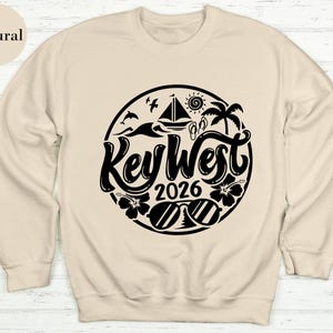 A beige sweatshirt with a black graphic design featuring the text "Key West" and "2026" surrounded by various beach-related icons such as a sailboat, palm tree, and sun.