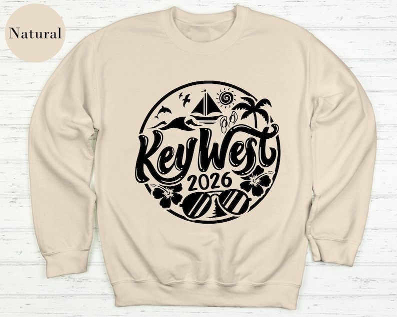 A beige sweatshirt with a black graphic design featuring the text "Key West" and "2026" surrounded by various beach-related icons such as a sailboat, palm tree, and sun.