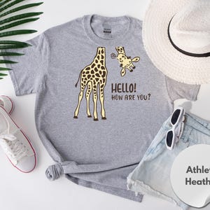 a gray t-shirt with a giraffe graphic and the text "Hello! How are you?" printed on it, along with a straw hat, sunglasses, and athletic shorts.