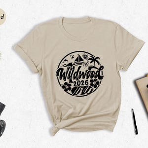 a beige t-shirt with a graphic design, a potted plant, a camera, a pen, a book, and a cup of coffee arranged on a white surface.