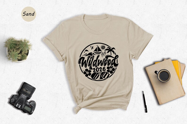 a beige t-shirt with a graphic design, a potted plant, a camera, a pen, a book, and a cup of coffee arranged on a white surface.