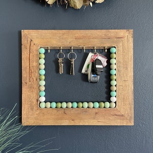 May include: A wooden frame with a beaded border in shades of green and white. The frame has hooks for hanging keys and other items. There are keys and a car key fob hanging from the hooks.