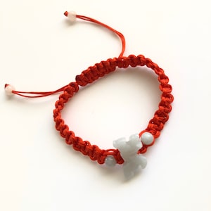 May include: A red string bracelet with a white jade charm in the shape of a bear. The bracelet is tied with a knot and has two white beads on the ends.