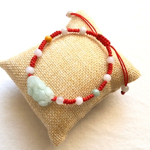 May include: A red braided bracelet with a jade pendant in the shape of a mythical creature. The bracelet features white and pink beads, and a yellow bead. The bracelet is displayed on a tan burlap pillow.