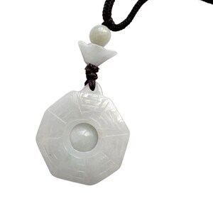 May include: A white jade pendant shaped as an octagon with a central circular cutout, suspended from a black braided cord. The pendant features engraved lines radiating from the center and is accented by a small white jade bead and a flower-shaped spacer.