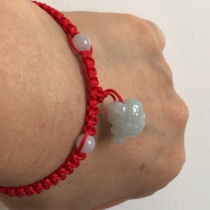 May include: Red braided bracelet with two light purple beads and a light green jade frog charm. The bracelet is tied with a sliding knot closure.