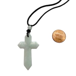 May include: A pale green stone cross pendant suspended on a black cord. The cross features faceted edges and a small bead at the top. A United States one-cent coin is placed next to the pendant to provide a sense of scale for the object's size.