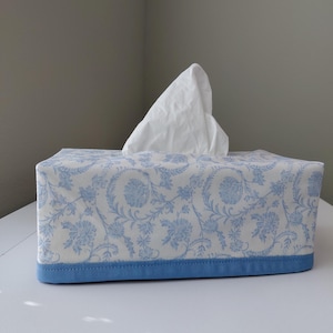 May include: A rectangular tissue box cover featuring a white fabric base with a light blue floral and vine pattern. The bottom edge is finished with a solid light blue fabric border. A single white tissue is pulled through the top opening.