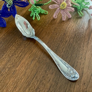 May include: A silver-toned metal spoon featuring a pointed, spade-shaped bowl and a handle decorated with embossed floral and leaf motifs. The handle tapers toward the bowl and displays intricate detailing at the base, showcasing a polished finish.