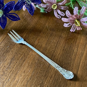 May include: A silver-colored, three-pronged fork with a decorative handle. The fork is resting on a wooden surface, with beaded flower decorations in the background. The fork is designed for serving or eating.