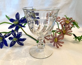 May include: A clear glass goblet with a fluted design and a short stem. The goblet is sitting on a white surface with blue and pink flowers.